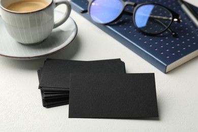 Photo of Blank black business cards, notebook, glasses and cup of coffee on white table, closeup. Mockup for design