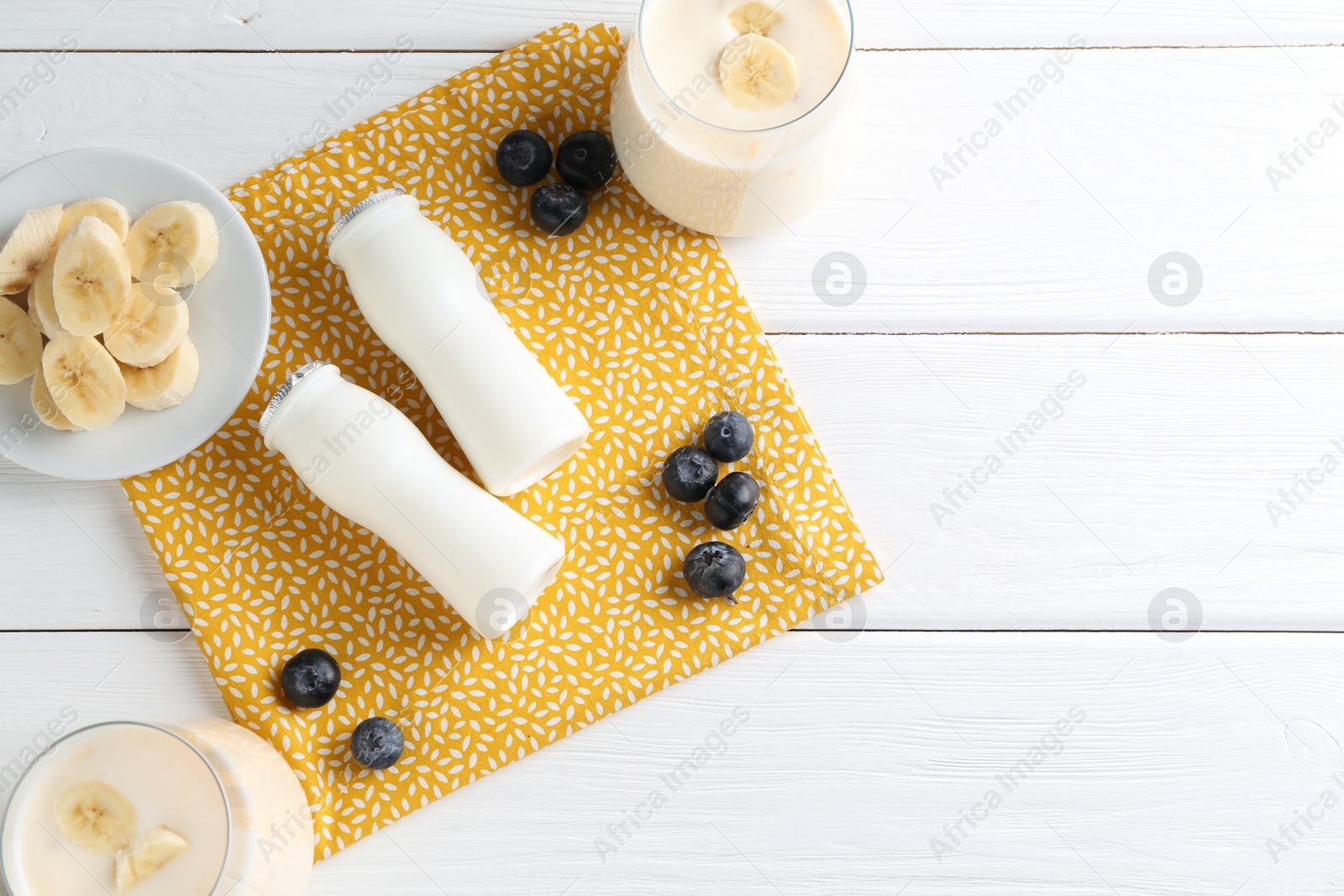 Photo of Tasty yogurt in bottles, banana and blueberries on white wooden table, top view. Space for text