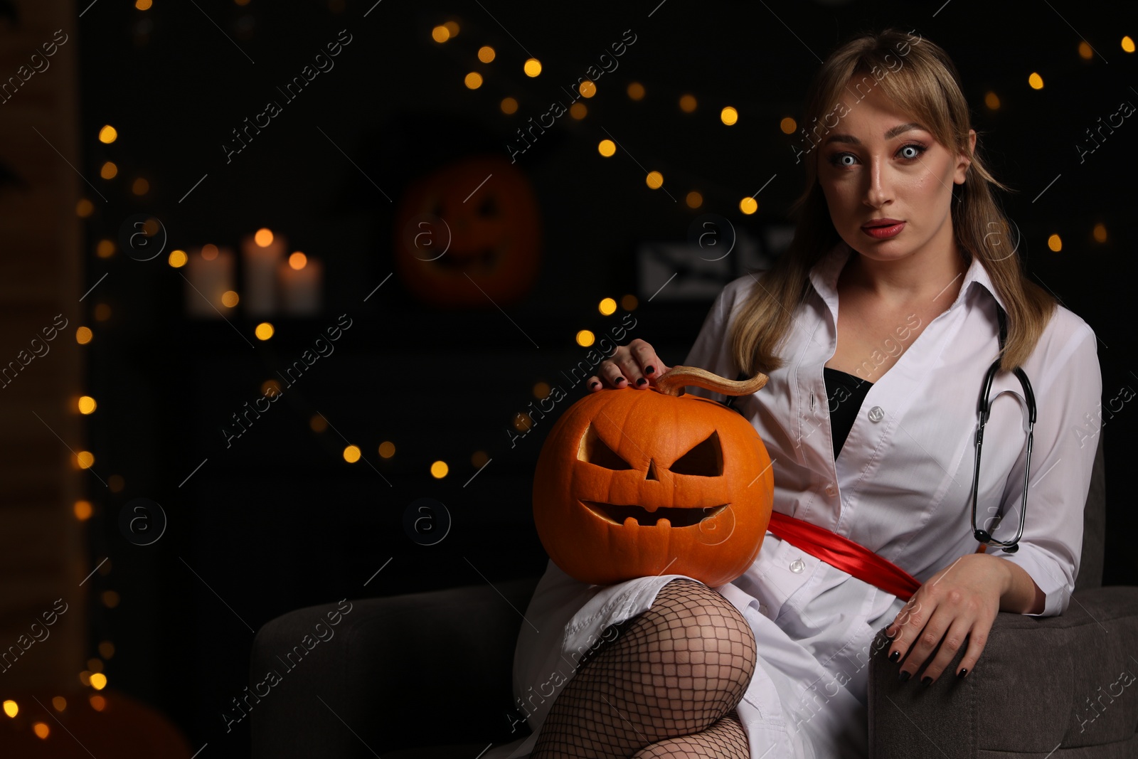 Photo of Woman in scary nurse costume with carved pumpkin against blurred lights indoors, space for text. Halloween celebration