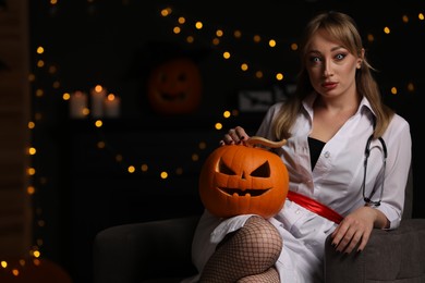 Woman in scary nurse costume with carved pumpkin against blurred lights indoors, space for text. Halloween celebration