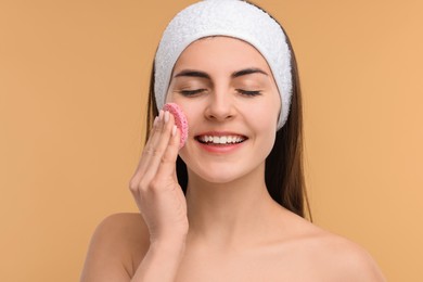 Photo of Young woman with headband washing her face using sponge on beige background