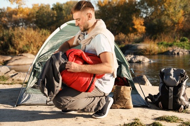 Photo of Young man packing sleeping bag near camping tent outdoors