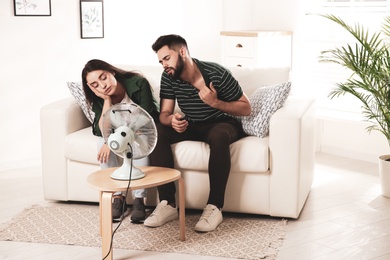 Photo of Couple suffering from heat in front of fan at home. Summer season