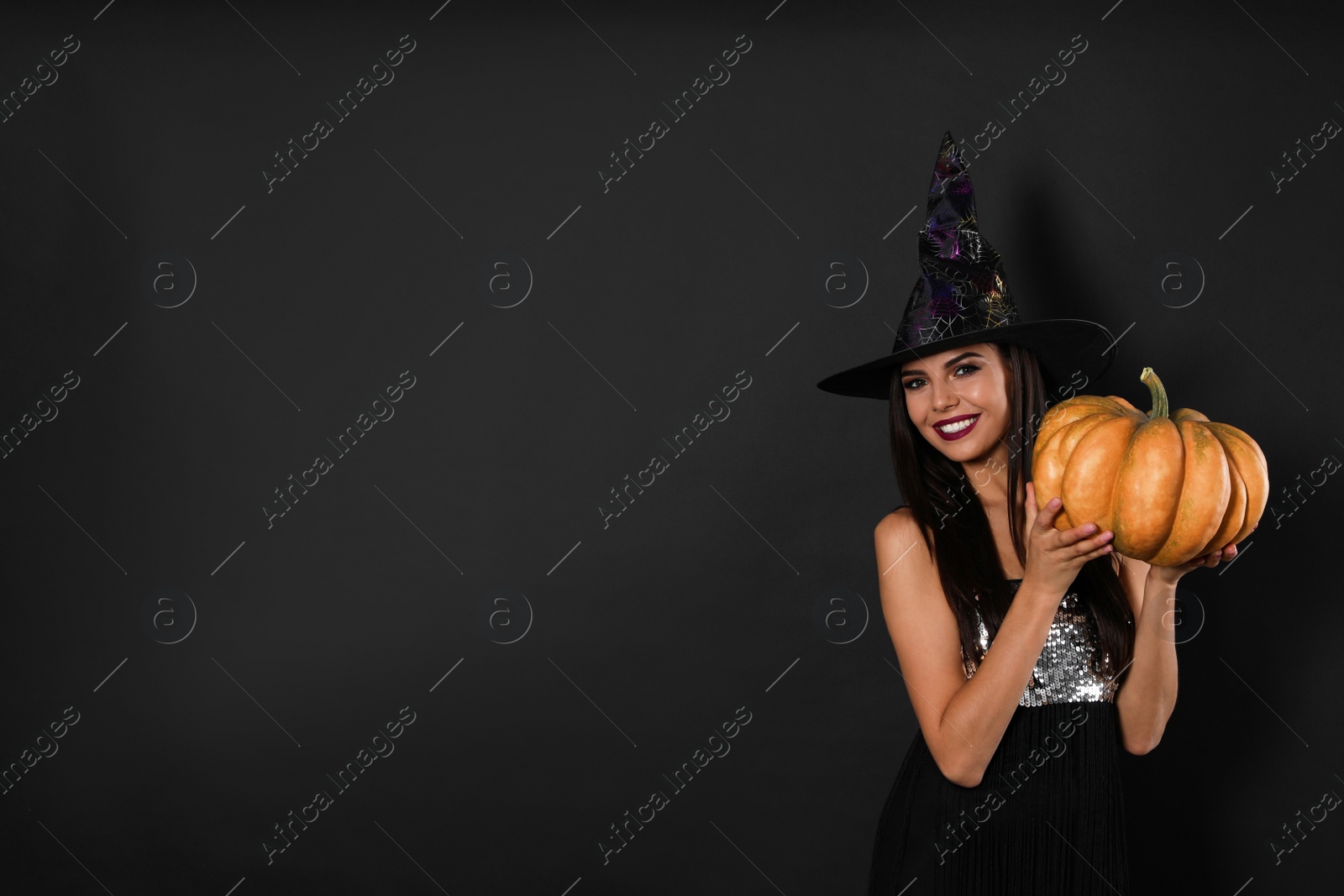 Photo of Beautiful woman wearing witch costume with pumpkin for Halloween party on black background, space for text