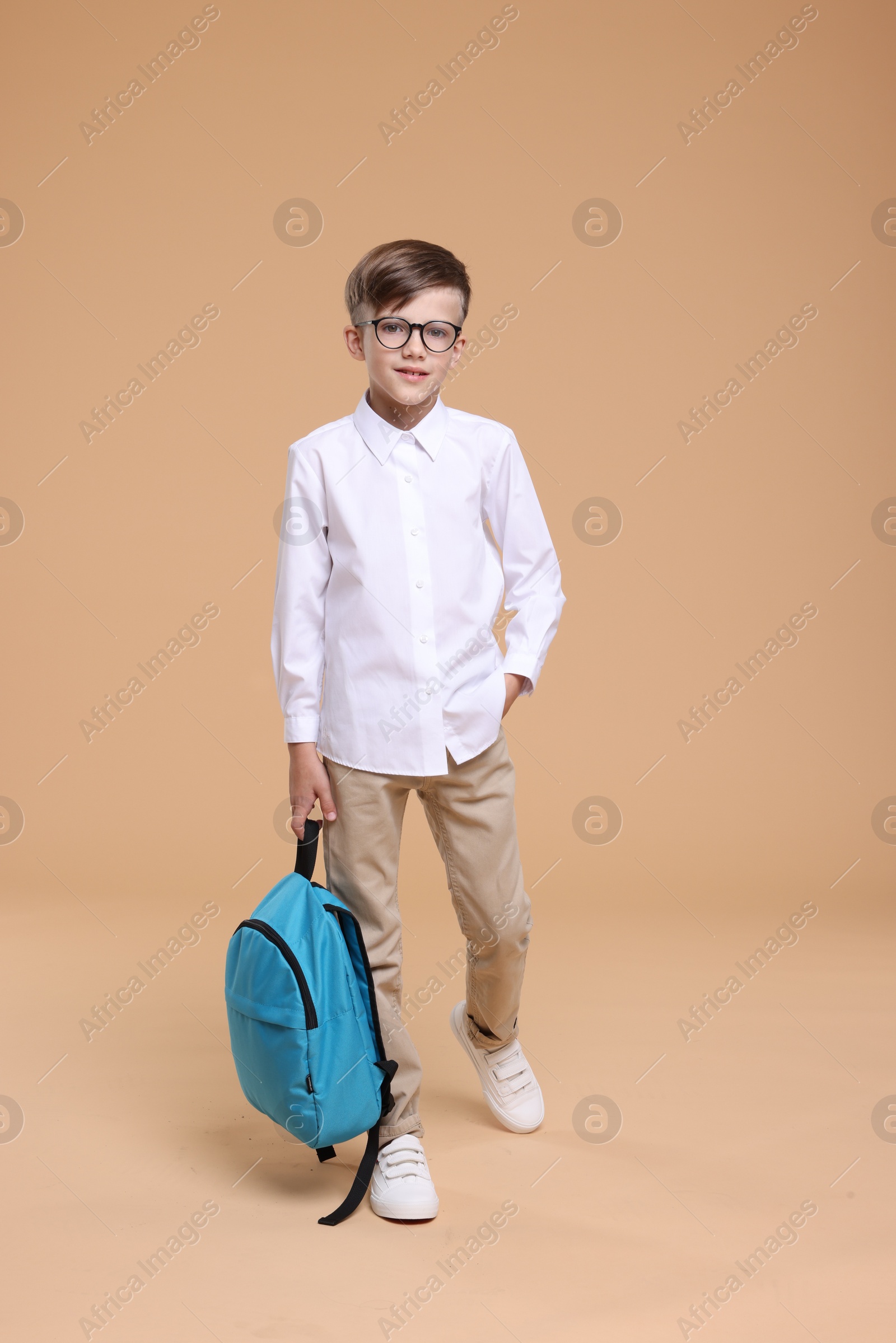 Photo of Cute schoolboy in glasses holding backpack on beige background