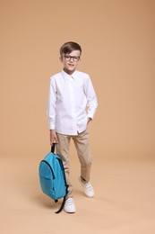 Cute schoolboy in glasses holding backpack on beige background