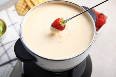 Dipping strawberry into pot with white chocolate fondue on table