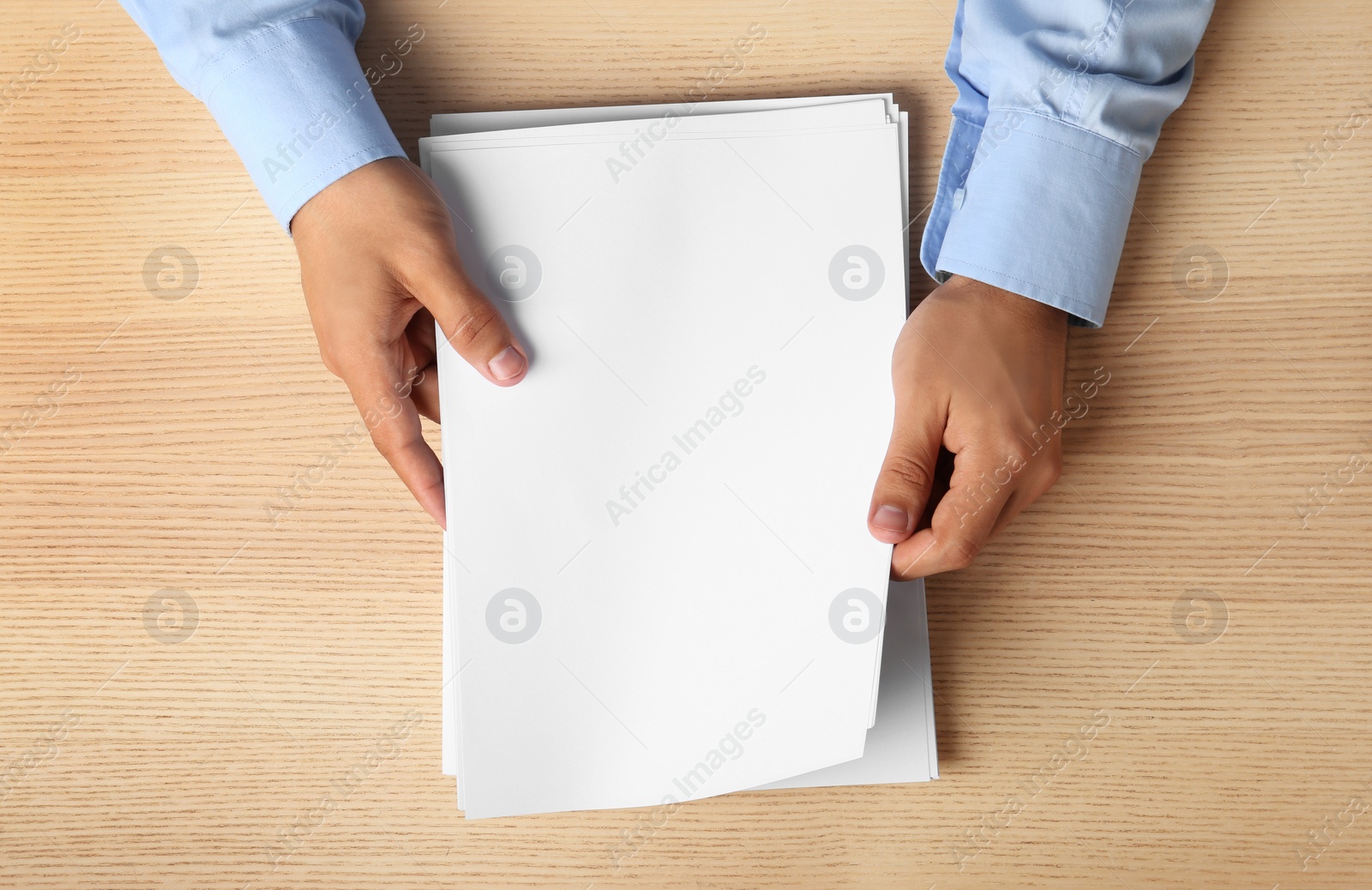 Photo of Man holding blank paper sheets for brochure at wooden table, top view. Mock up