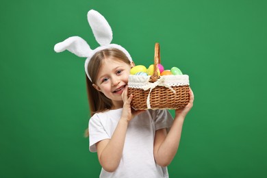 Photo of Easter celebration. Cute girl with bunny ears holding basket of painted eggs on green background