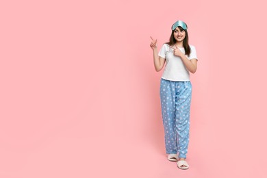 Happy woman in pyjama and sleep mask pointing at something on pink background, space for text