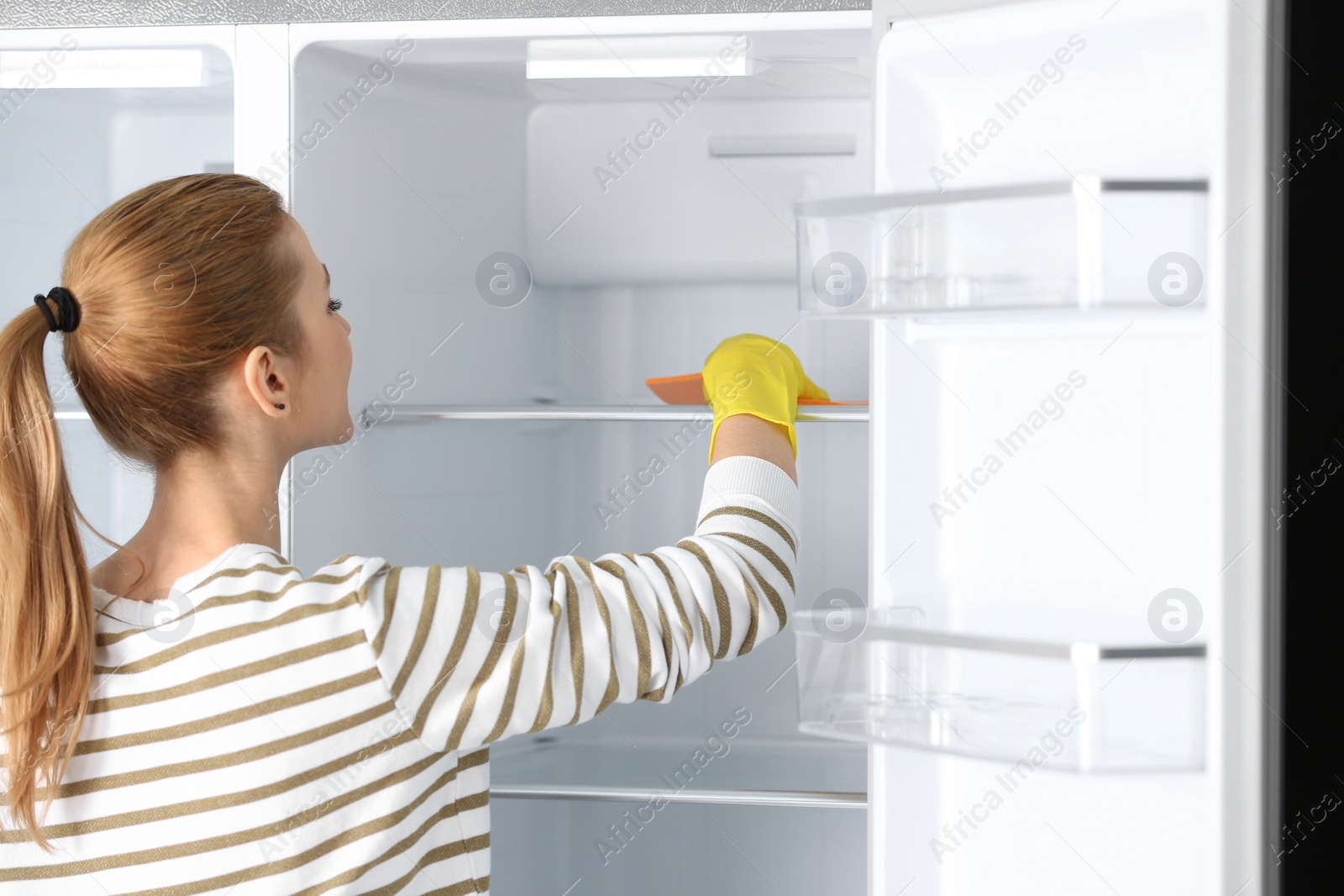 Photo of Woman in rubber gloves cleaning empty refrigerator with rag at home