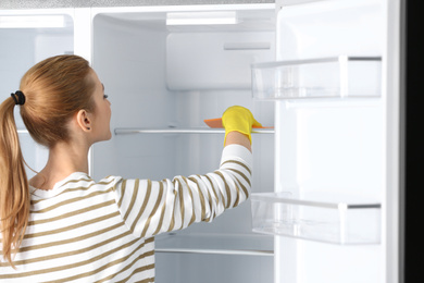 Photo of Woman in rubber gloves cleaning empty refrigerator with rag at home