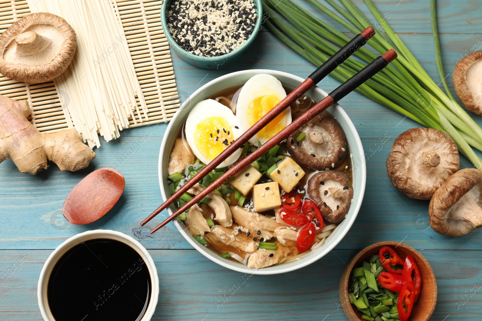 Photo of Bowl of delicious ramen and ingredients on light blue wooden table, flat lay. Noodle soup