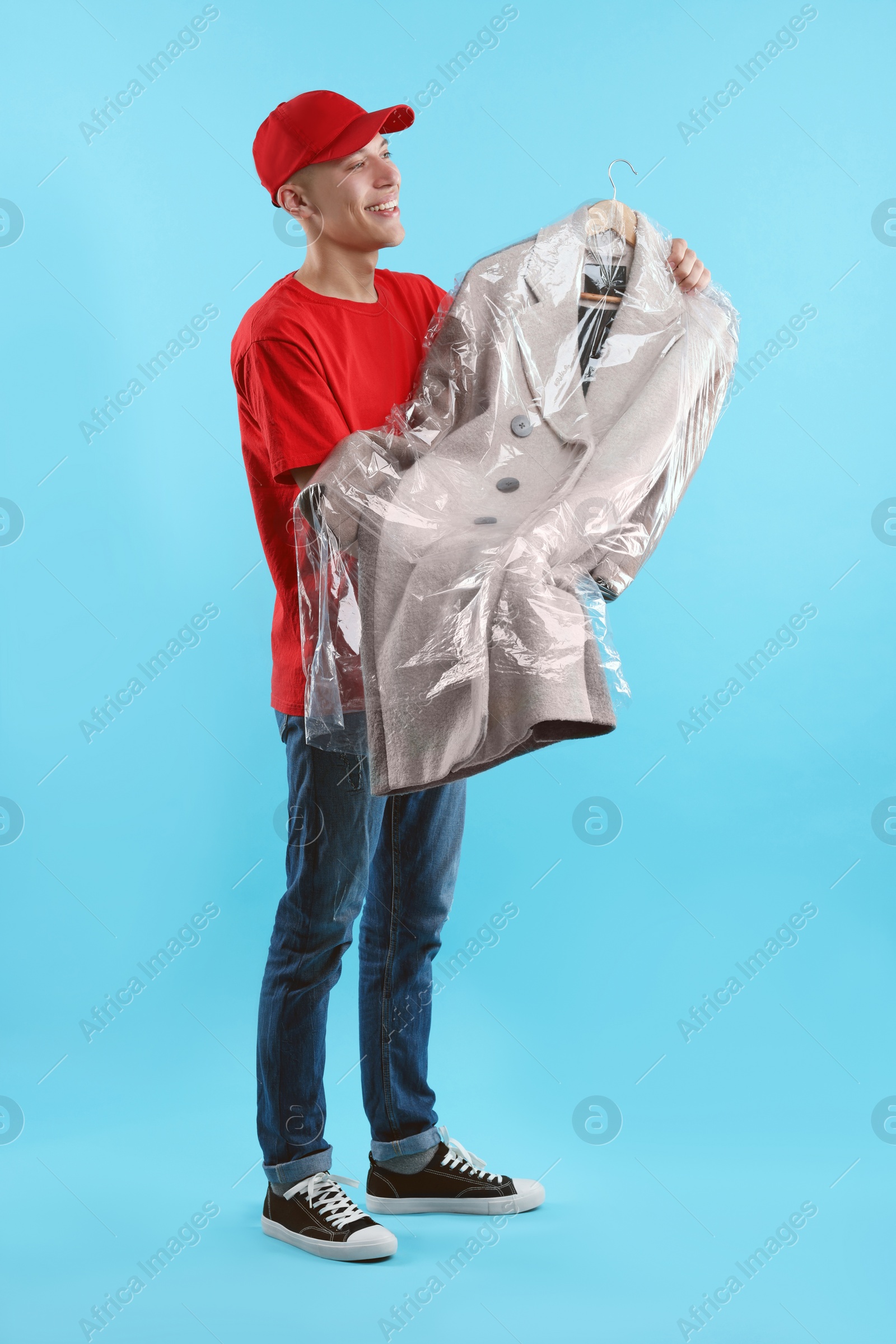 Photo of Dry-cleaning delivery. Happy courier holding coat in plastic bag on light blue background