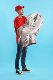 Photo of Dry-cleaning delivery. Happy courier holding coat in plastic bag on light blue background