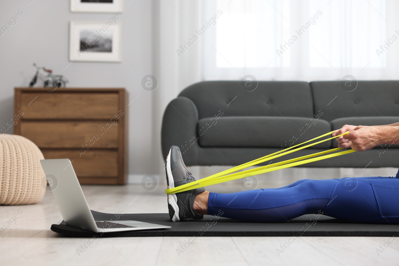 Photo of Woman doing exercise with fitness elastic band near laptop on mat at home, closeup