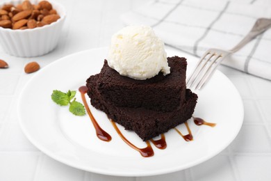 Photo of Tasty brownies served with ice cream and caramel sauce on white tiled table, closeup