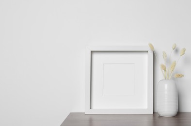 Photo of Empty photo frame and vase with dry decorative spikes on wooden table. Mockup for design