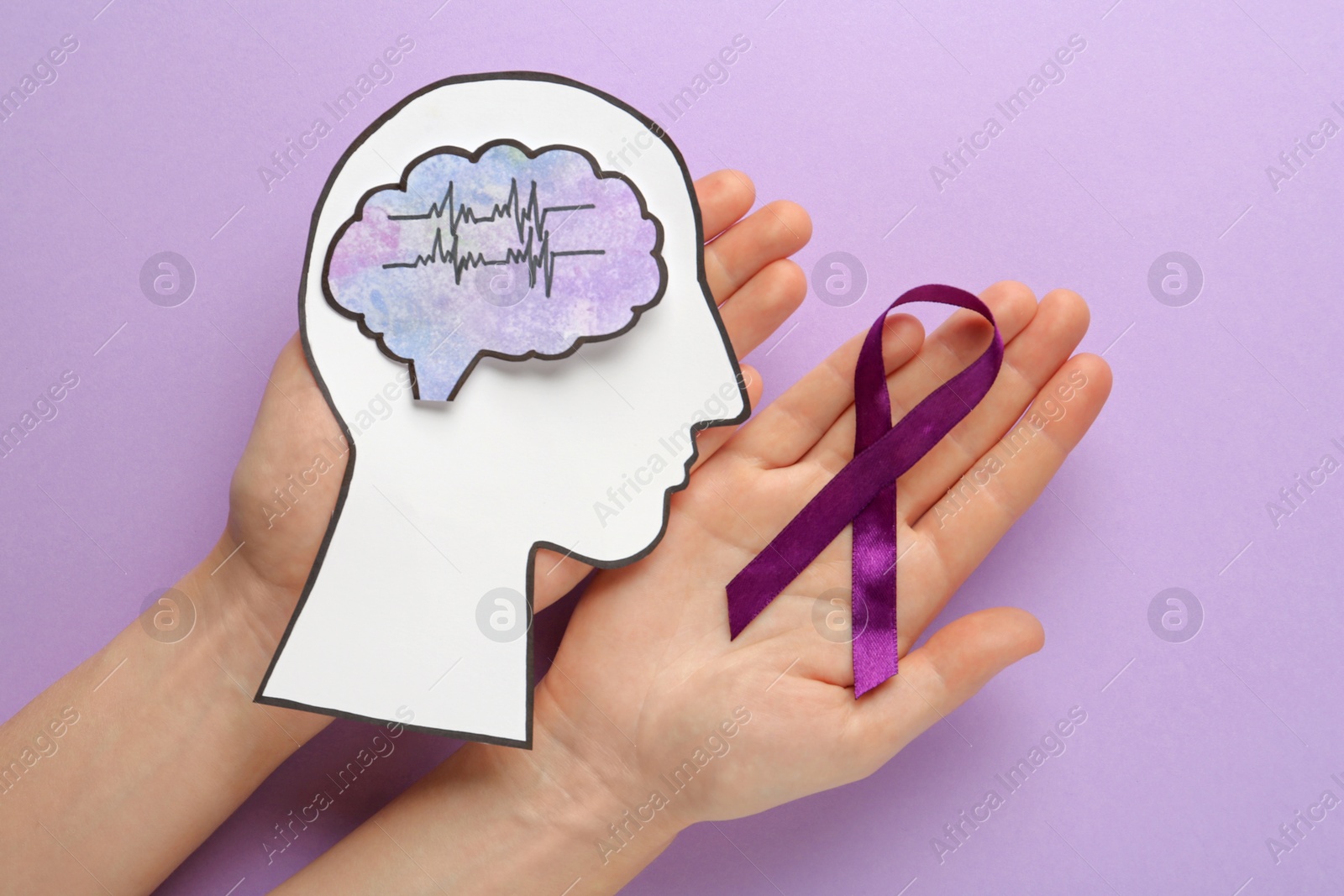 Photo of Woman holding human head cutout, brain and purple ribbon at violet background, top view. Epilepsy awareness