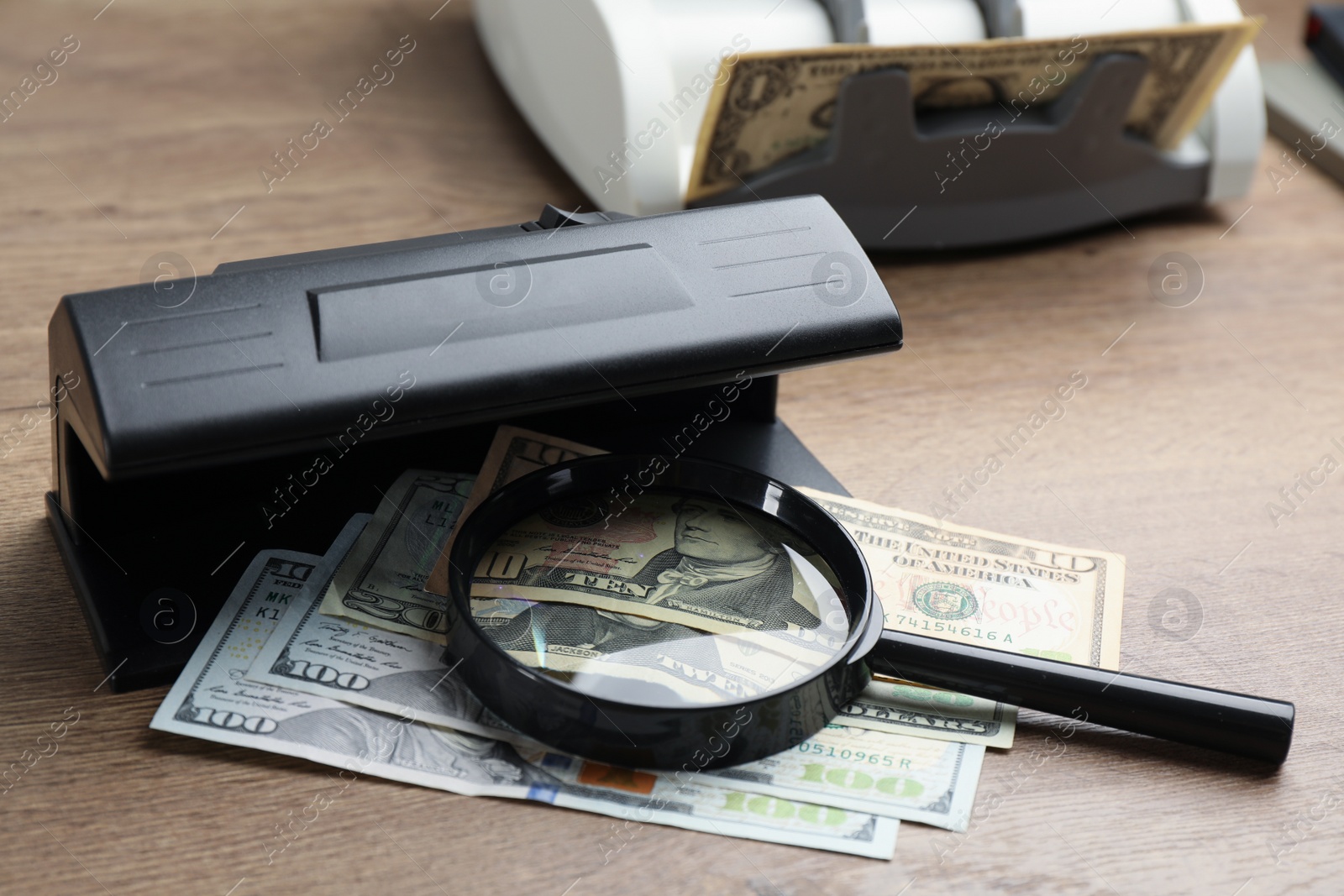 Photo of Modern currency detector with dollar banknotes and magnifying glass on wooden table. Money examination device