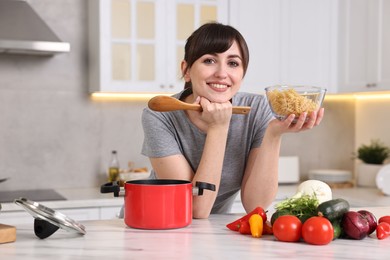 Photo of Happy young housewife with spoon and raw pasta at white marble table in kitchen. Cooking process