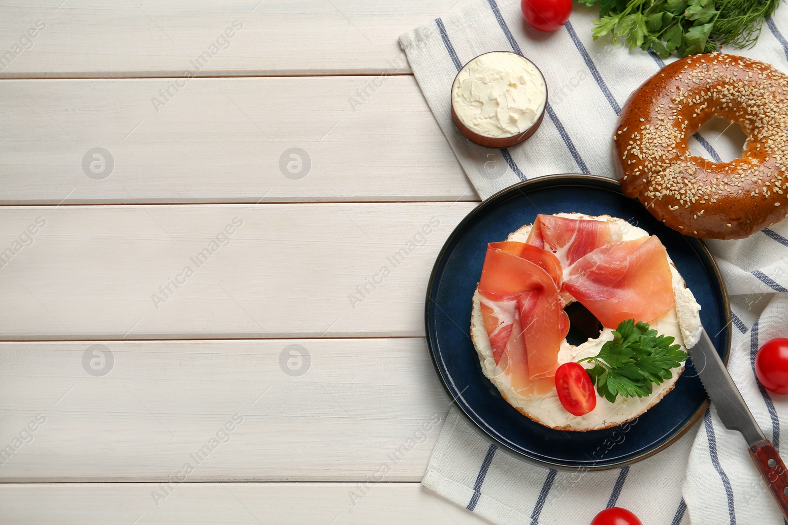 Photo of Delicious bagel with cream cheese, jamon, tomato and parsley on white wooden table, flat lay. Space for text