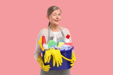 Happy housewife holding bucket with cleaning supplies on pink background