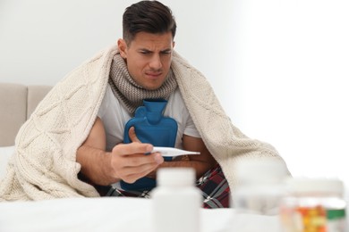 Photo of Ill man with hot water bottle checking his temperature at home