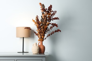 Photo of Stylish vases, dried eucalyptus branches and table lamp on chest near white wall indoors, space for text. Interior design