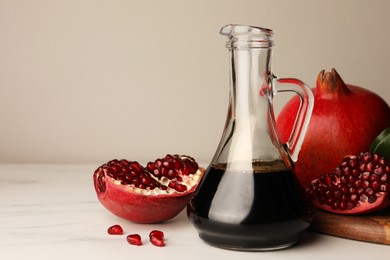 Photo of Glass jug of tasty pomegranate sauce and fresh ripe fruits on white marble table. Space for text