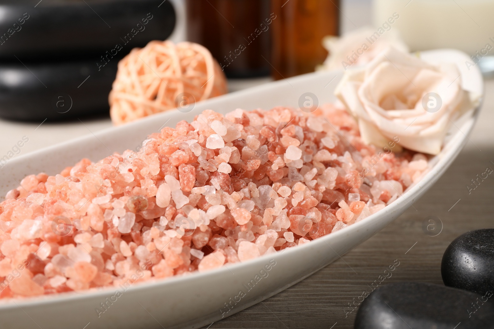 Photo of Natural sea salt in bowl, spa stones, rose and rattan ball on wooden table, closeup