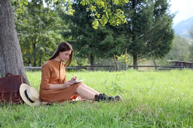 Photo of Beautiful young woman drawing with pencil in notepad near tree on green grass