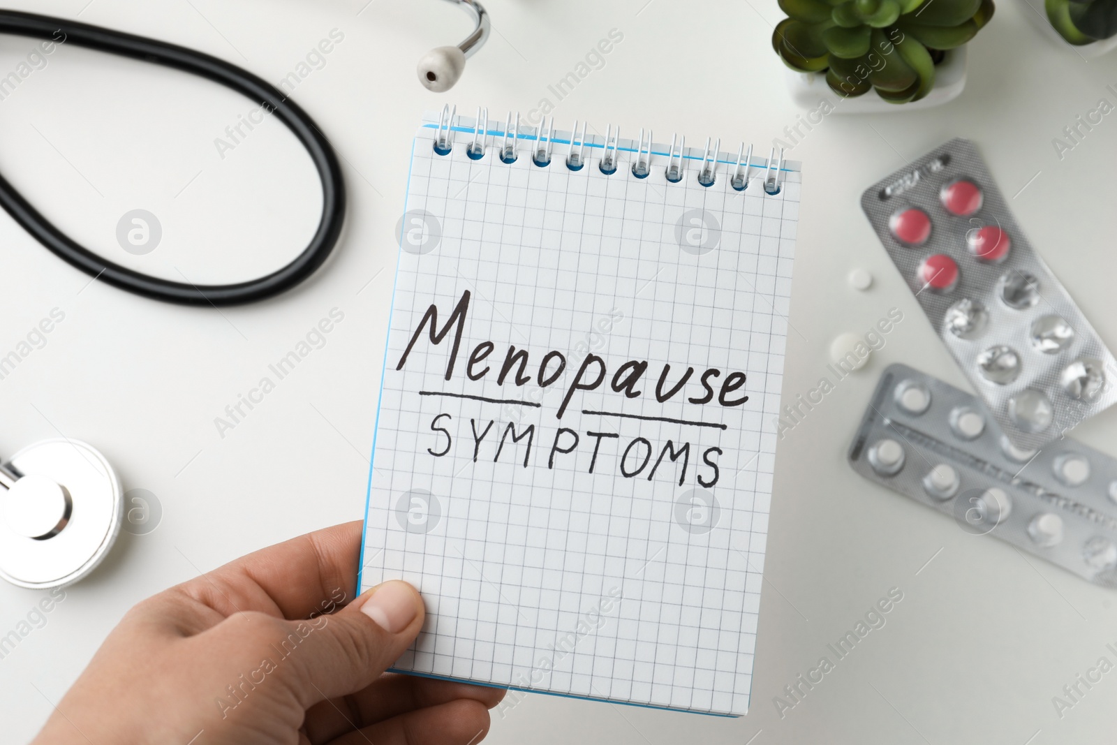 Photo of Woman holding notebook with words Menopause Symptoms at white table, top view