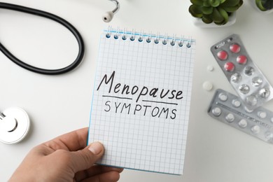 Woman holding notebook with words Menopause Symptoms at white table, top view