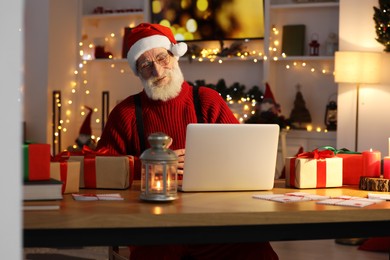 Photo of Santa Claus using laptop and smiling at his workplace in room decorated for Christmas