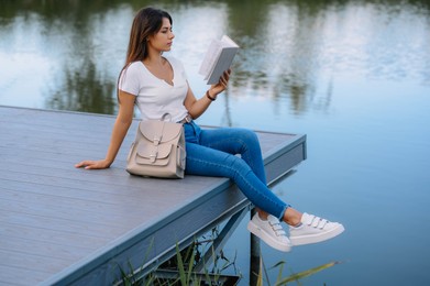 Photo of Young woman reading book on pier near lake