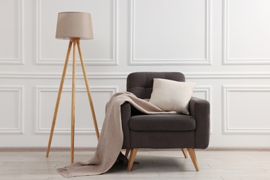 Photo of Stylish armchair with cushion, blanket and lamp near white wall indoors