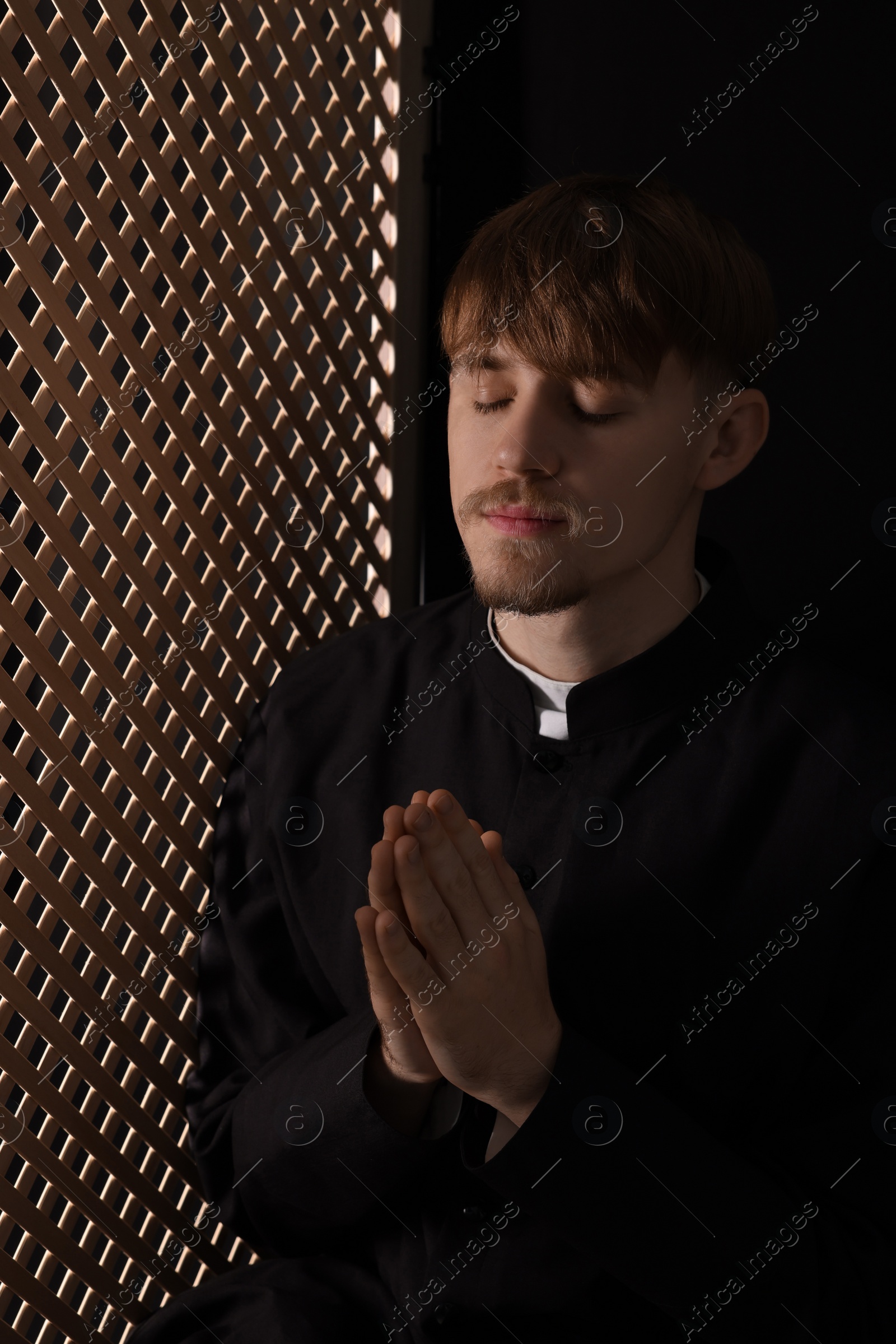 Photo of Catholic priest praying near wooden window in confessional booth