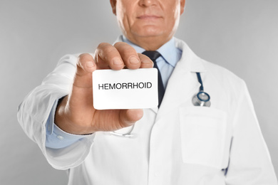 Photo of Doctor holding business card with word HEMORRHOID on light grey background, closeup