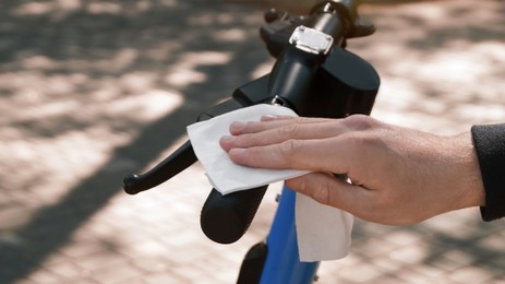 Photo of Man cleaning scooter steering wheel with wet wipe outdoors, closeup. Protective measures