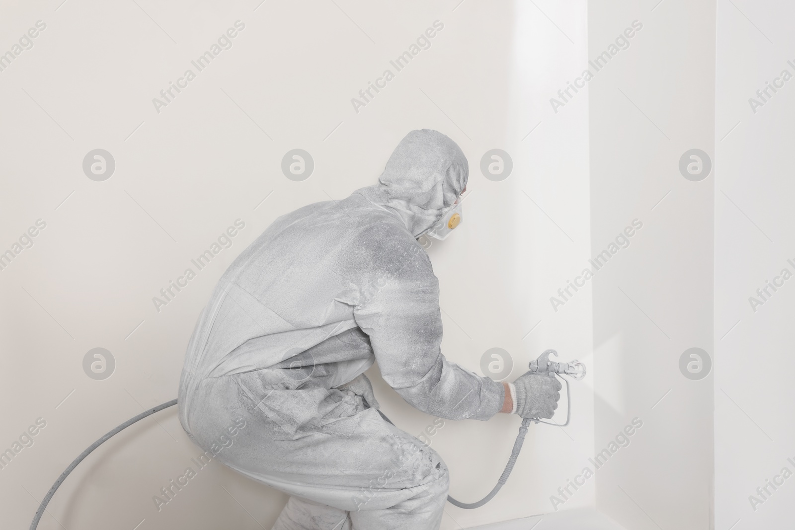 Photo of Decorator in protective overalls painting wall with spray gun indoors