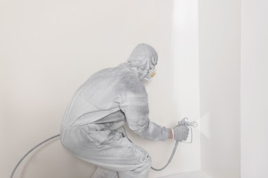 Decorator in protective overalls painting wall with spray gun indoors