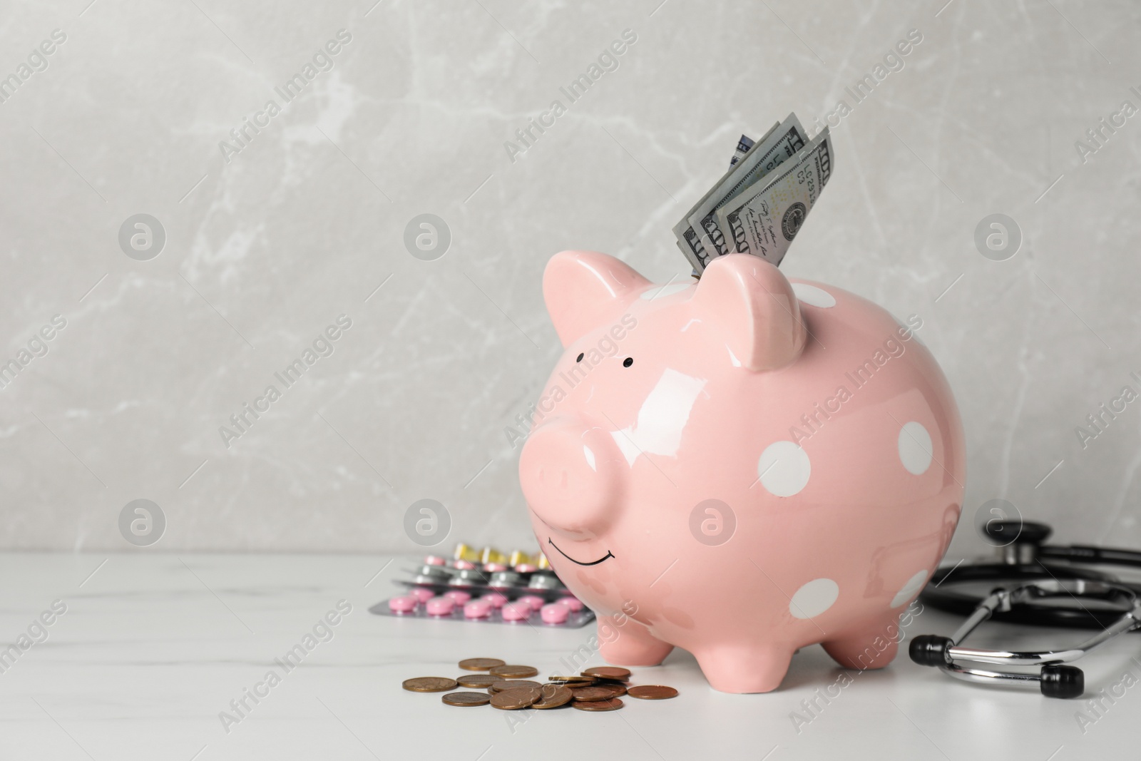 Photo of Pale pink ceramic piggy bank with money, stethoscope and pills on white table, space for text. Medical insurance