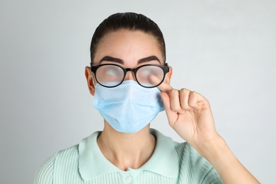 Woman wiping foggy glasses caused by wearing medical mask on light background