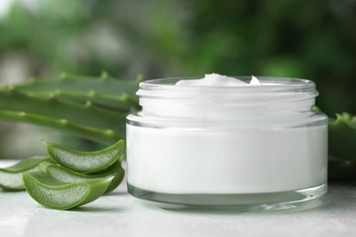 Photo of Jar with cream and cut aloe leaves on white table against blurred green background, closeup