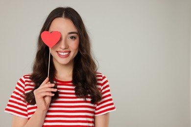 Photo of Beautiful young woman covering her eye with paper heart on grey background, space for text