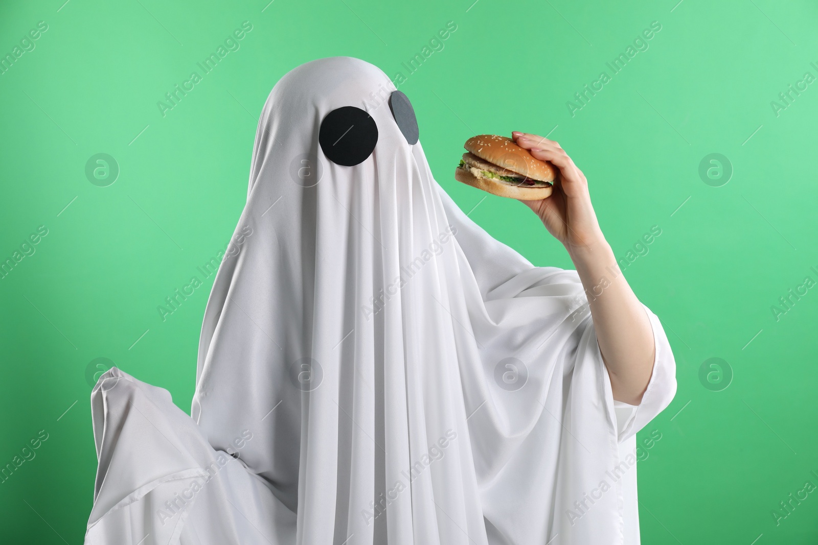 Photo of Creepy ghost. Person in white sheet with tasty hamburger on green background