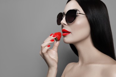 Image of Attractive woman in stylish sunglasses holding strawberry on grey background, space for text. Color accent