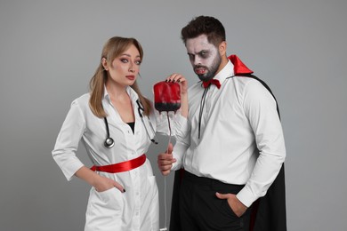 Couple in scary nurse and vampire costumes with blood bag on light grey background. Halloween celebration