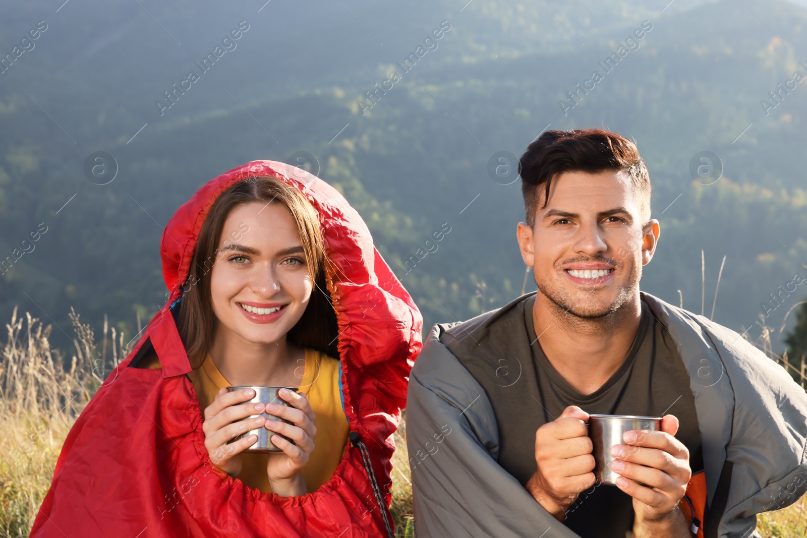 Photo of Happy couple with drinks in sleeping bags surrounded by beautiful nature